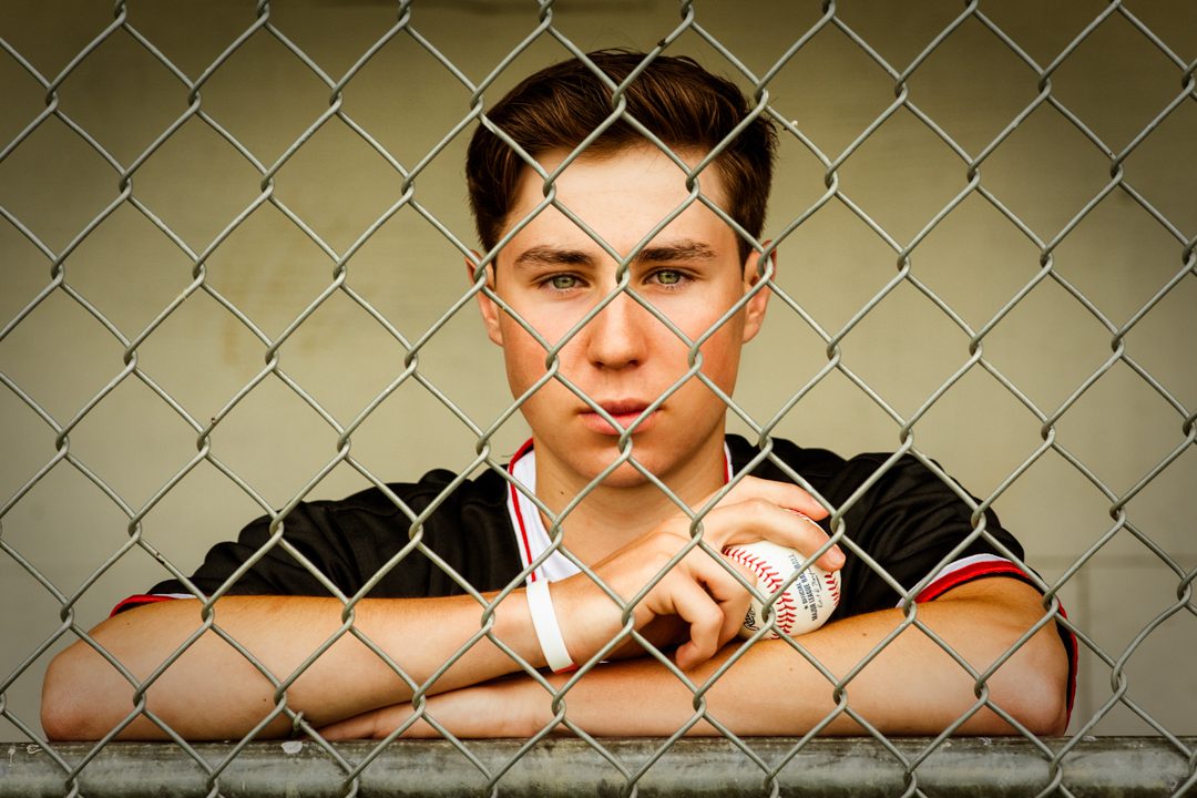 A young man leaning against the fence of his baseball dugout.