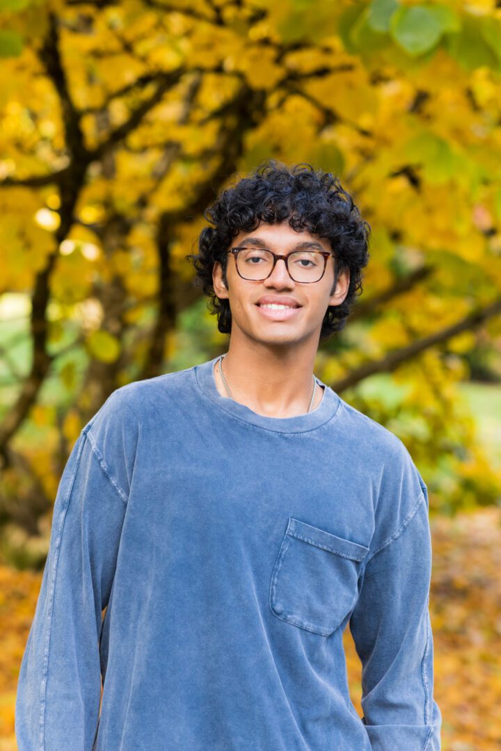 A young man with glasses standing in front of trees.