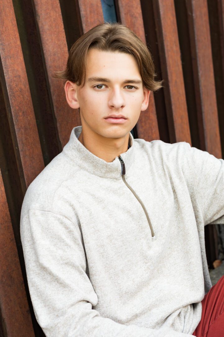 A young man in grey sweater standing next to wooden fence.