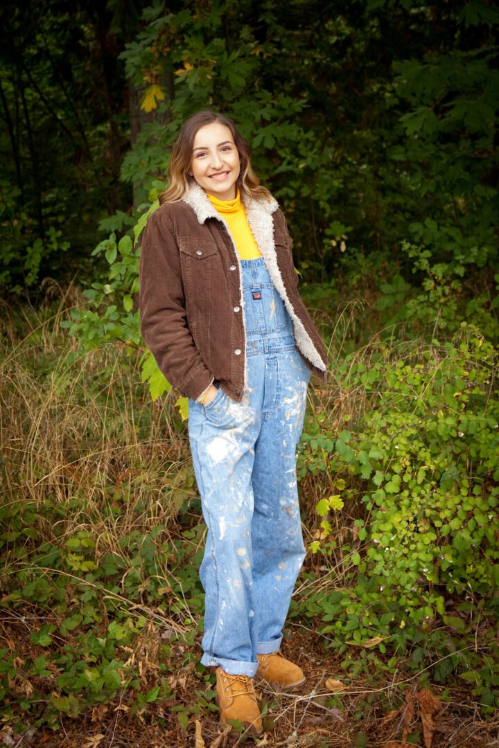 A woman standing in the woods wearing overalls.